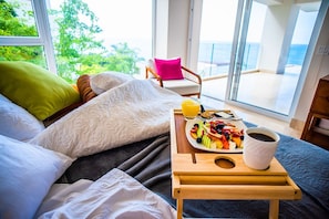 A bedroom with a bed, a breakfast tray with fruits, a cup of coffee, and a view of the sea through large windows.