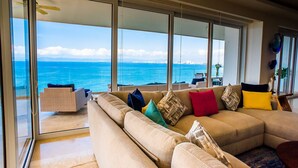 Un salon doté d’une grande fenêtre donnant sur l’océan, d’un canapé beige agrémenté de coussins colorés et d’une porte en verre qui mène à un espace extérieur.