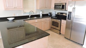 A kitchen with light-colored cabinets, stainless steel appliances, a granite countertop, and a window with blinds.