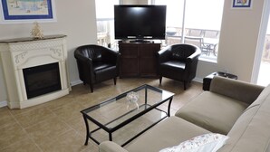 A living room with a fireplace, two leather armchairs, a glass coffee table, a beige sofa, a television, and a painting on the wall.