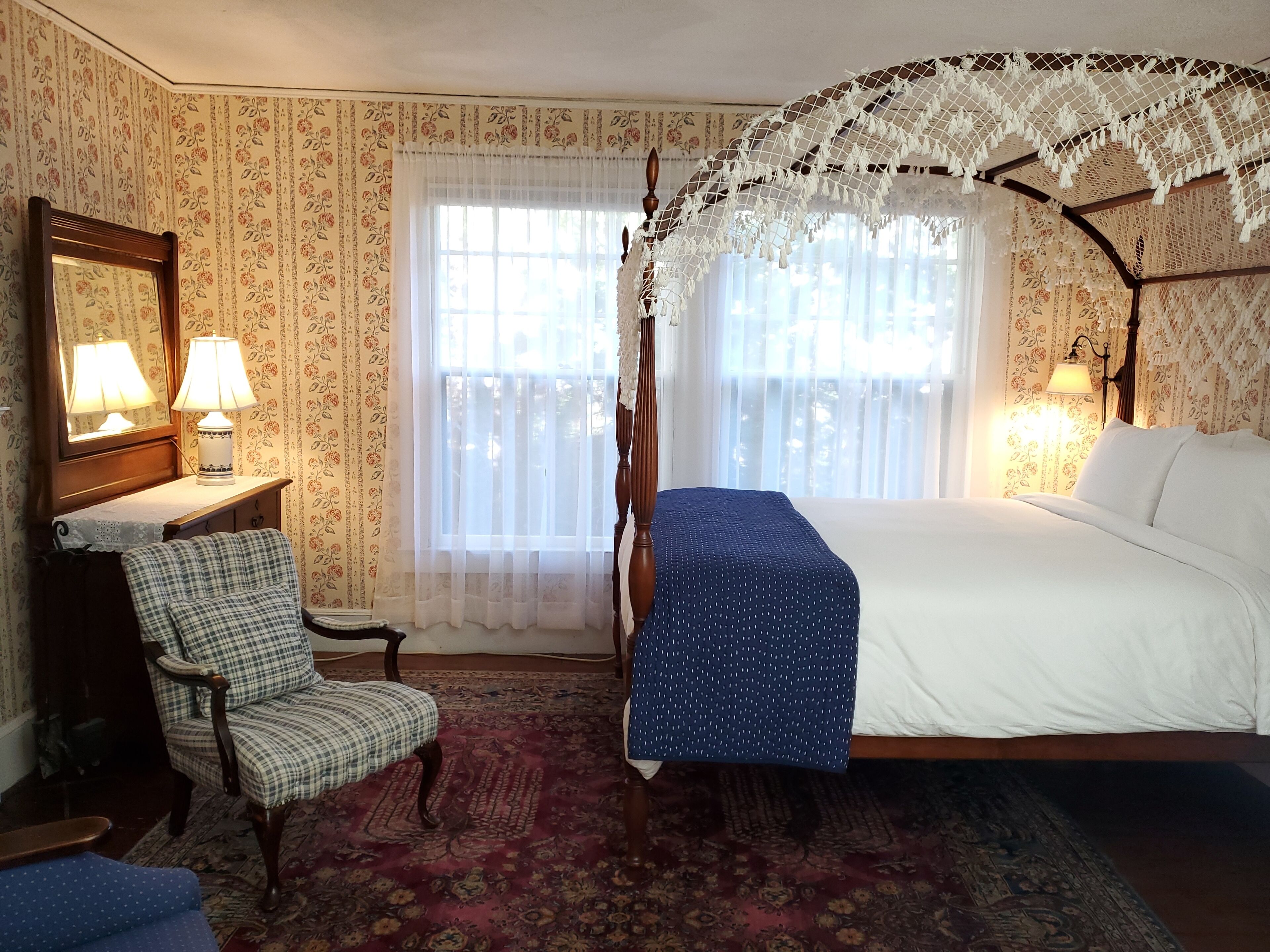 Un dormitorio con una cama con dosel, una silla, una mesita de noche con lámparas y una ventana con cortinas de encaje.