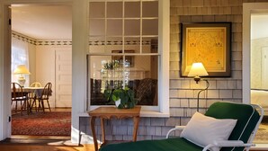 A cozy room with a green lounge chair, a wooden table with a lamp, a framed map on the wall, and a view of a dining area. - Harbourside Inn (Northeast Harbor)