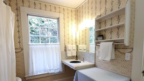 A bathroom with a wooden floor, a window with curtains, a sink, a mirror, shelves, and a towel rack.