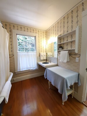 A bathroom with a wooden floor, a window with curtains, a sink, a mirror, shelves, and a towel rack.