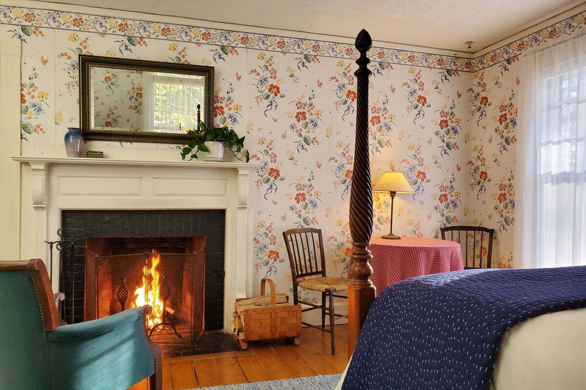 Una habitación con chimenea, papel tapiz floral, una cama, un sillón y una mesa con lámpara.