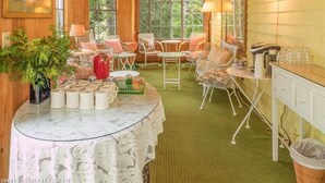 A cozy cabin interior with a dining area, chairs, a table with a lace tablecloth, and a view of the forest through large windows. - Harbourside Inn (Northeast Harbor)