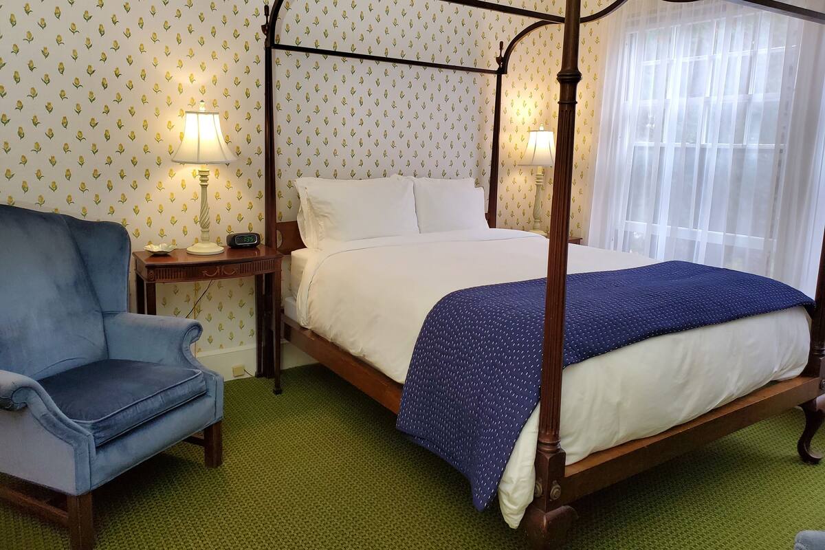 Una cama con dosel con sábanas blancas y una manta azul, un sillón azul y una mesita de noche con una lámpara.