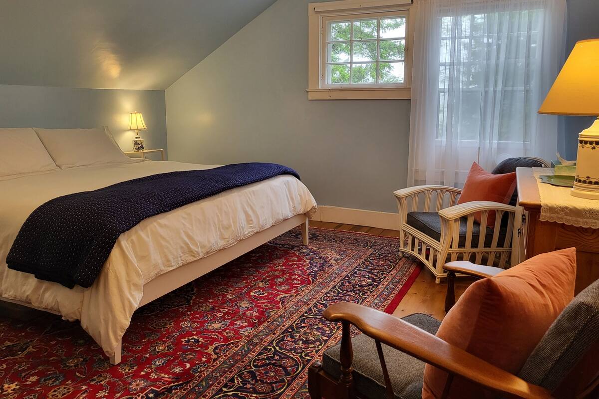 Un dormitorio con cama, una silla, una mesa, una lámpara y una ventana con cortinas.