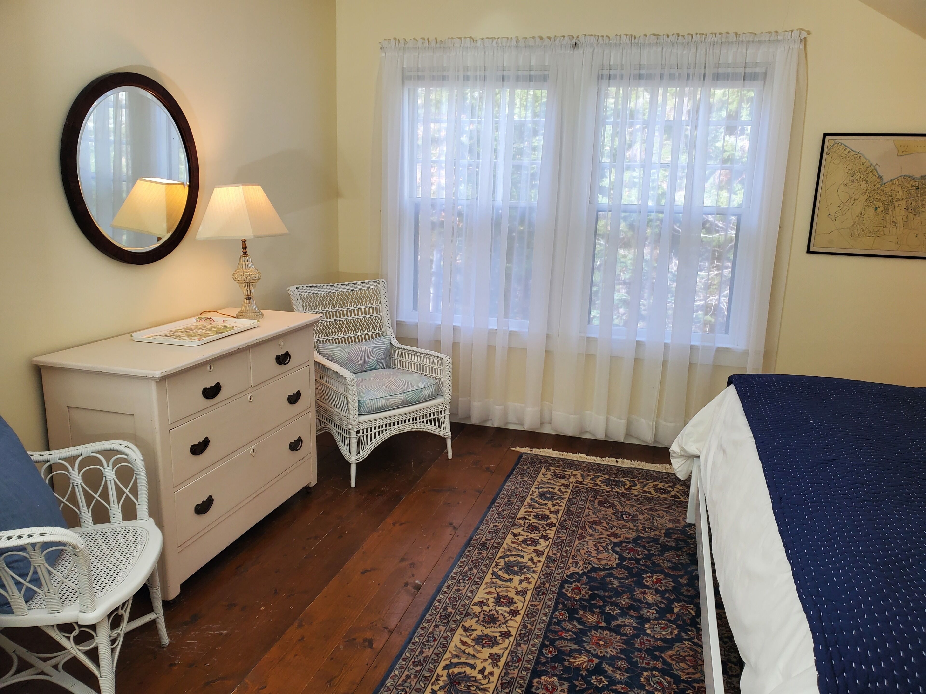 A bedroom with a wooden floor, a white dresser, a wicker chair, a bed with a blue and white bedspread, a large window with sheer curtains, and a wall-mounted mirror.