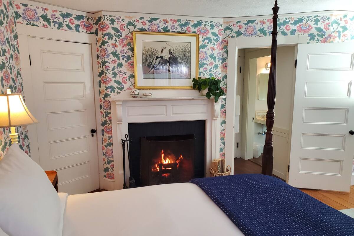 Un dormitorio con chimenea, papel tapiz floral, una cama con manta azul y una mesita de noche con lámpara.
