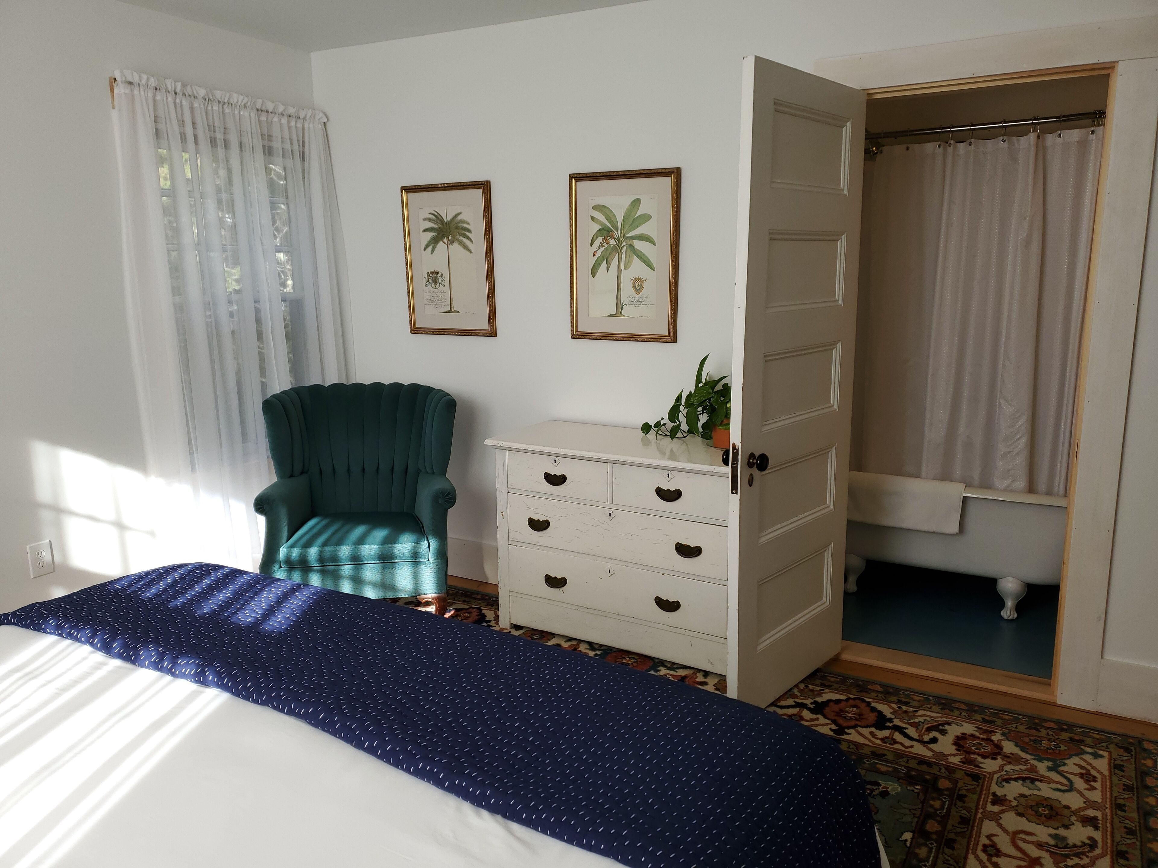 A bedroom with a bed, a green armchair, a white dresser with three drawers, and two framed pictures on the wall.
