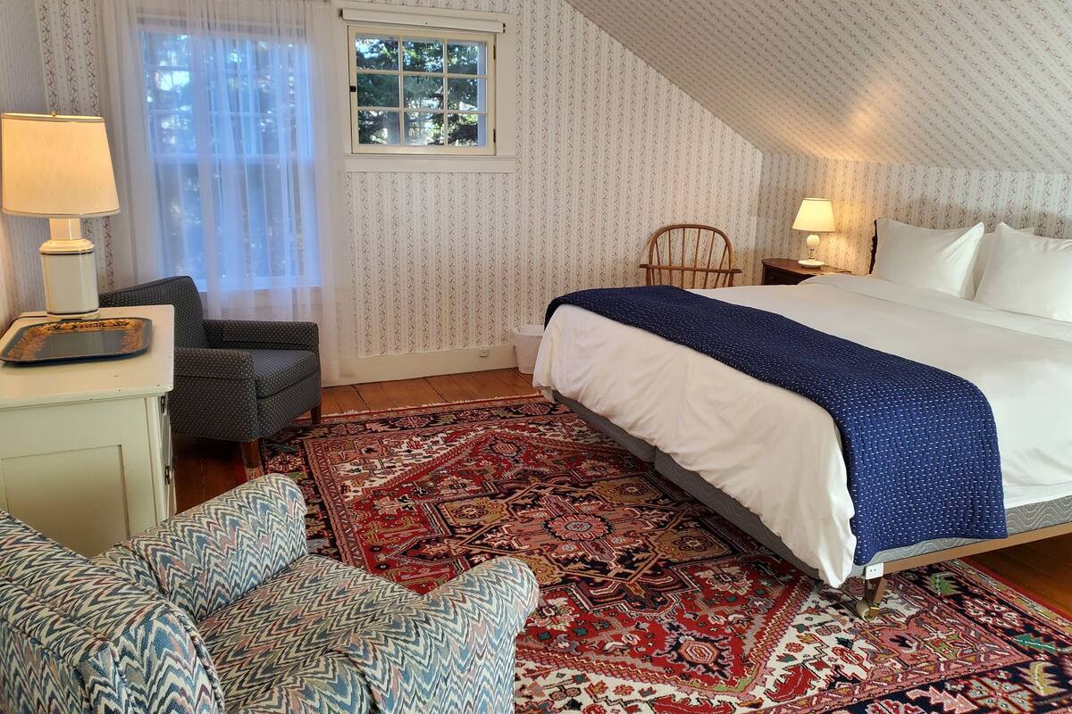 Un dormitorio con una cama grande, una silla, una mesita de noche, una lámpara, un ventanal y una alfombra estampada.