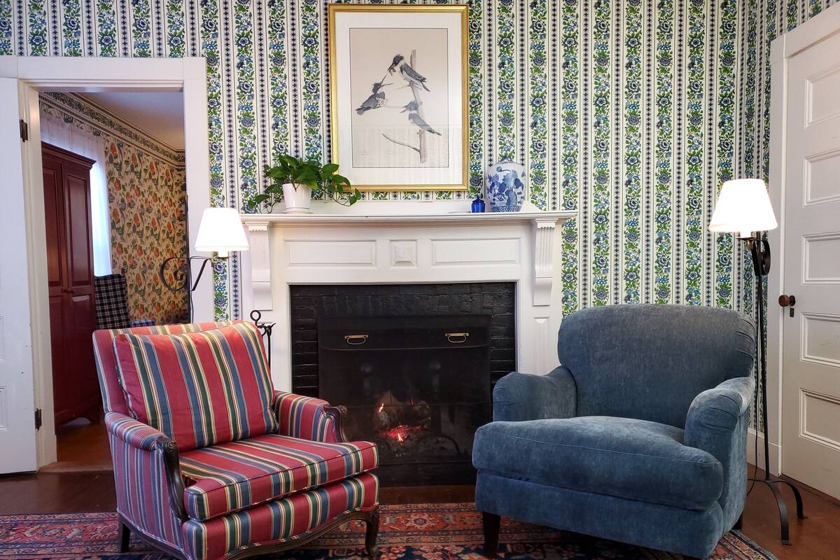 Una habitación con chimenea, un sillón a rayas, un sillón azul y papel tapiz estampado.