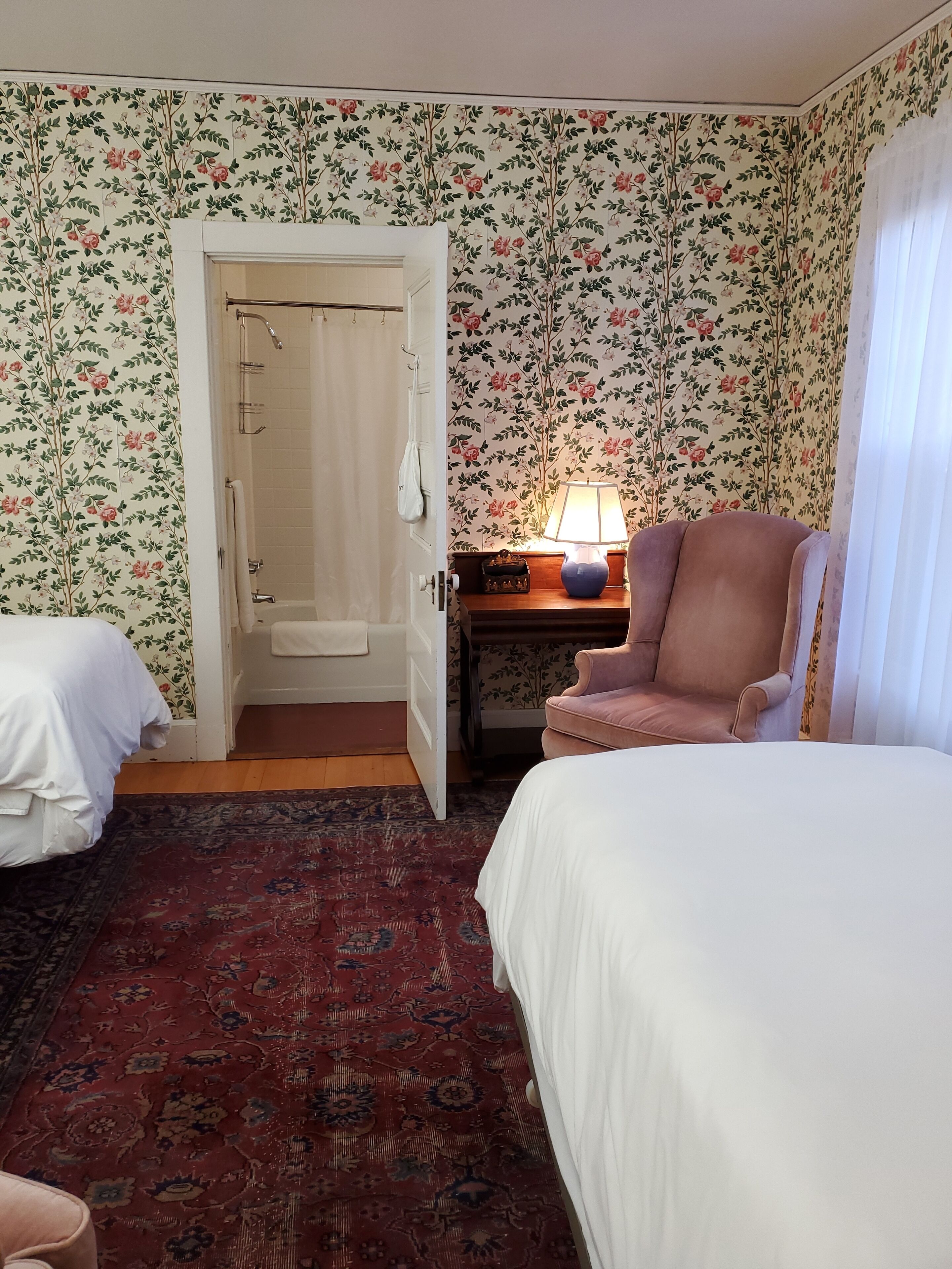 Una habitación con papel tapiz floral, una cama, una silla, una mesita de noche con lámpara y una puerta que da al baño.
