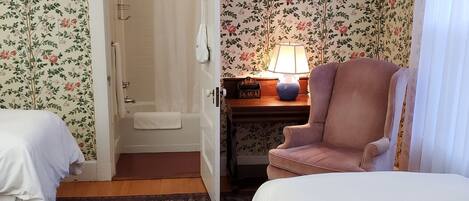 Una habitación con papel tapiz floral, una cama, una silla, una mesita de noche con lámpara y una puerta que da al baño.