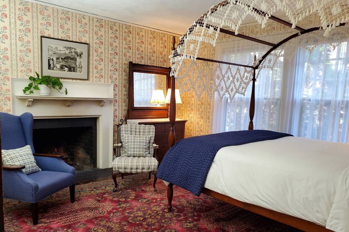 Una habitación con una cama con dosel, un sillón azul, un sillón a cuadros, una chimenea y una alfombra estampada.