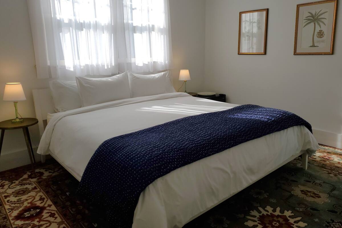 Una cama bien hecha con sábanas blancas y una manta azul, una lámpara de noche y un cuadro en la pared.