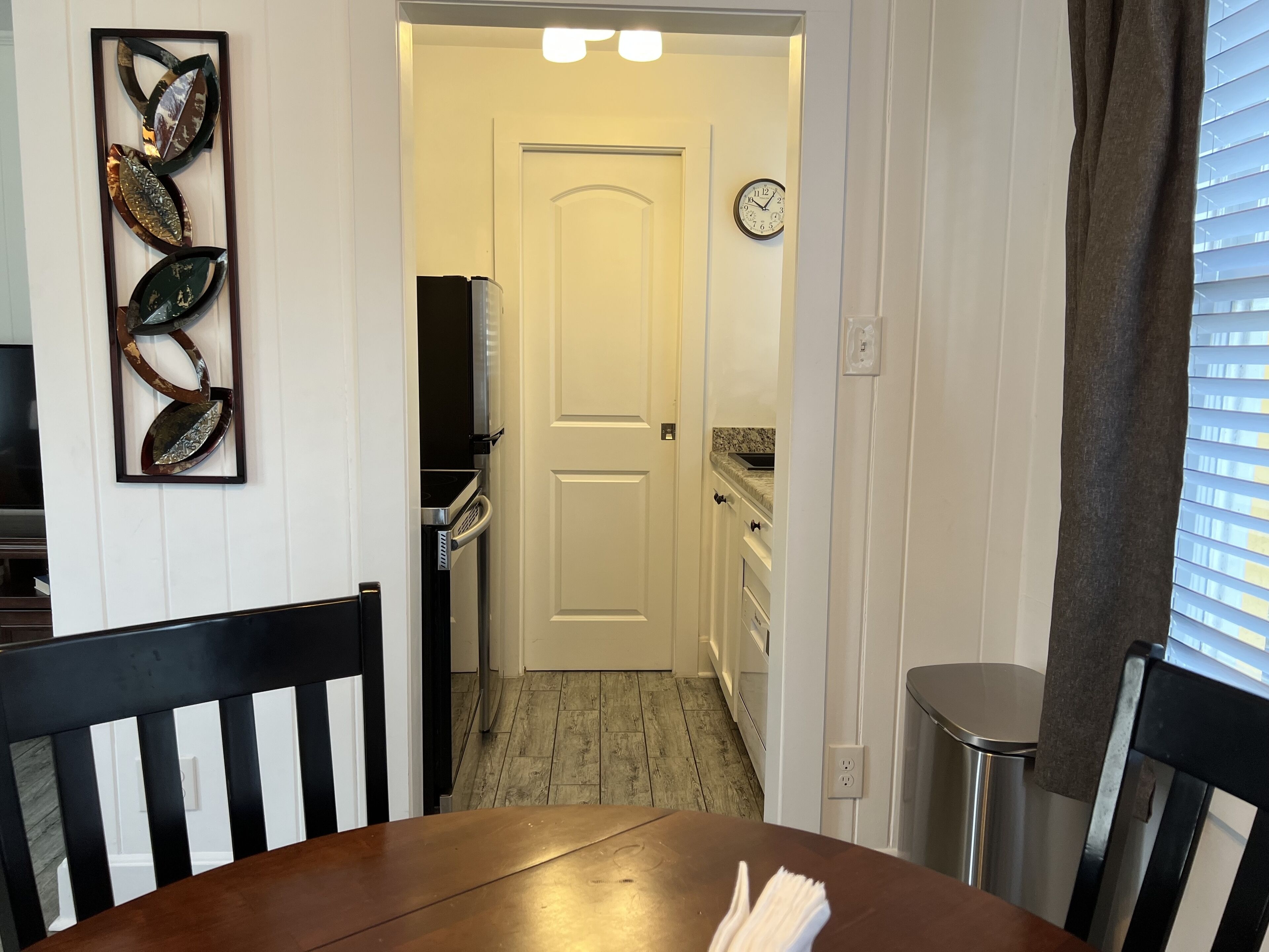 A dining area with a wooden table and chairs, a kitchen in the background, and a clock on the wall.