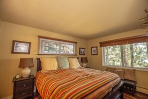 Quarto com uma cama grande, duas mesas de cabeceira com luminárias, um banquinho ao pé da cama e uma janela com persianas.