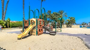 Un parque infantil con tobogán y estructuras de escalada, rodeado de palmeras y un cielo azul despejado.