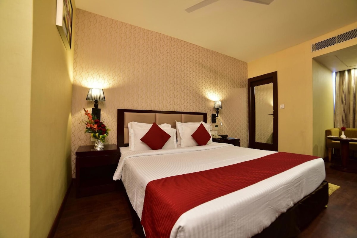 A hotel room with a large bed, two bedside lamps, a nightstand with a vase of flowers, a mirror, and a view of a dining area.