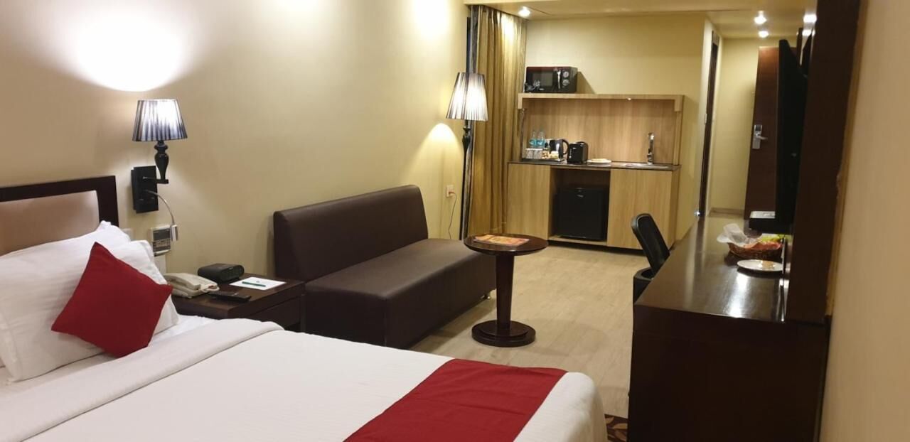 A hotel room with a bed, a sofa, a small table, a desk, a mirror, and a kitchenette in the background.