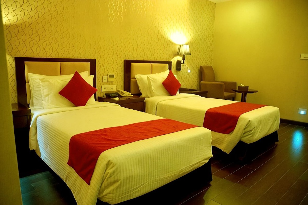 A hotel room with two beds, each with a red accent pillow, a bedside table with a lamp, and a chair.