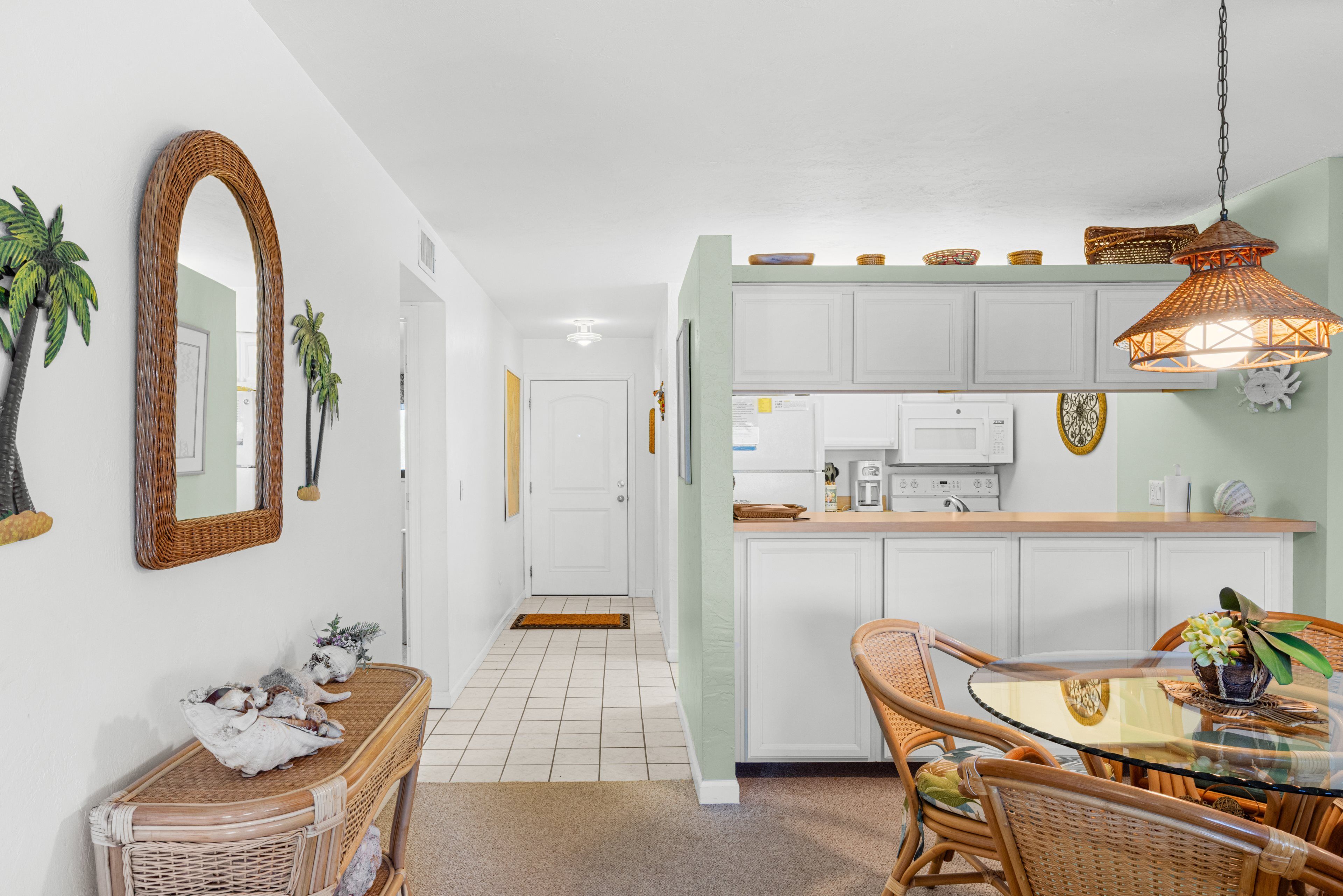 A modern kitchen with white cabinets, a dining area with wicker furniture, and a hallway with a white door.