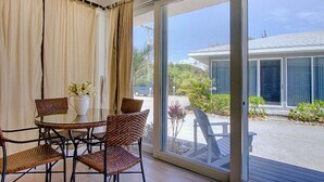 Una habitación con puerta de vidrio que da a un espacio exterior cubierto, sillas de mimbre, una mesa pequeña y un florero con flores.