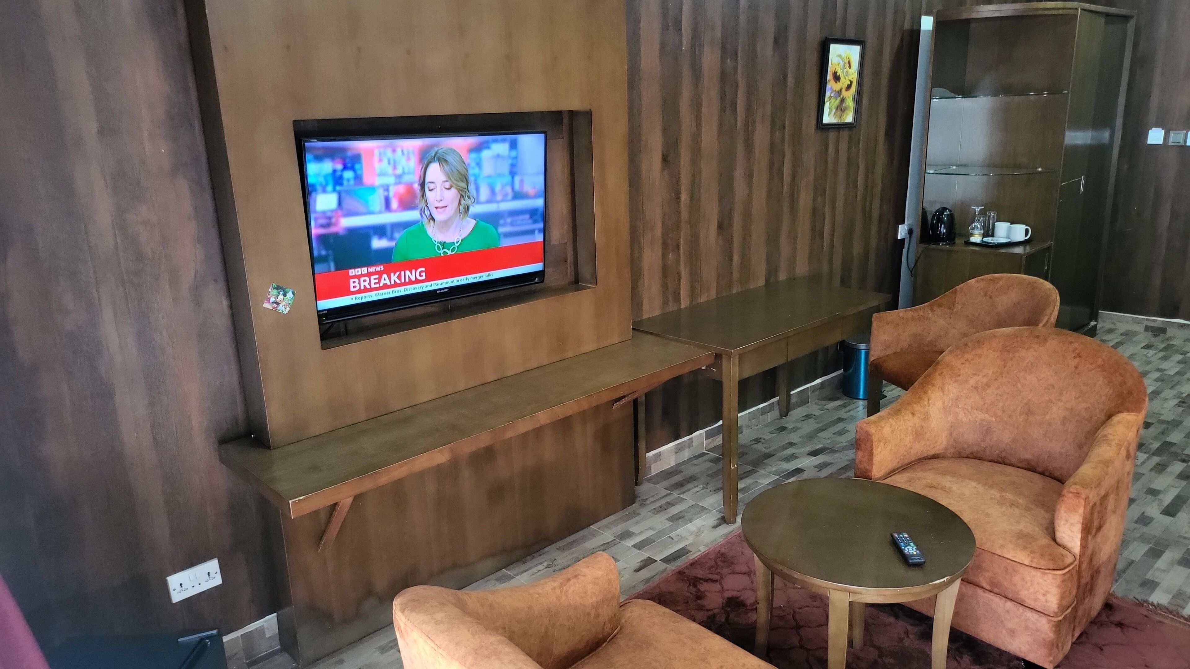 A modern living room with a flat-screen TV mounted on a wooden wall, a wooden bench, two plush armchairs, and a small round table.