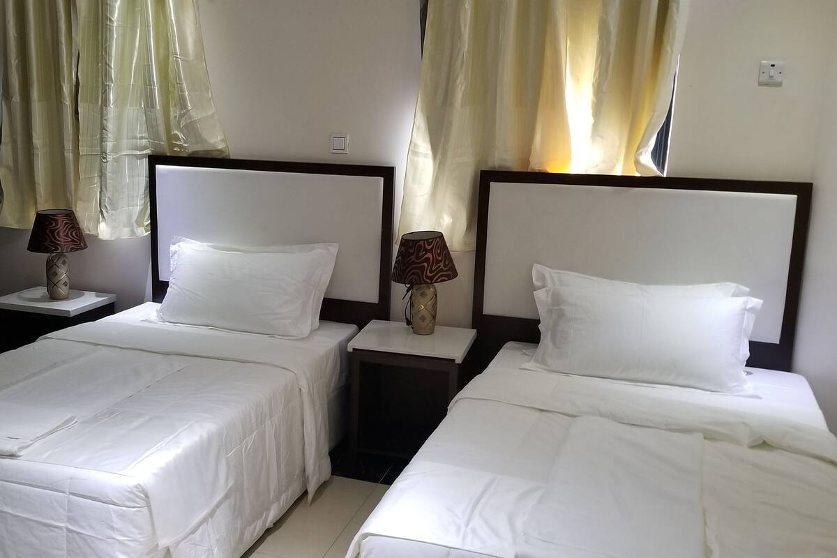 A hotel room with two single beds, each with a white bedspread and matching pillows. There are two bedside tables with lamps, and the room has a door and curtains.