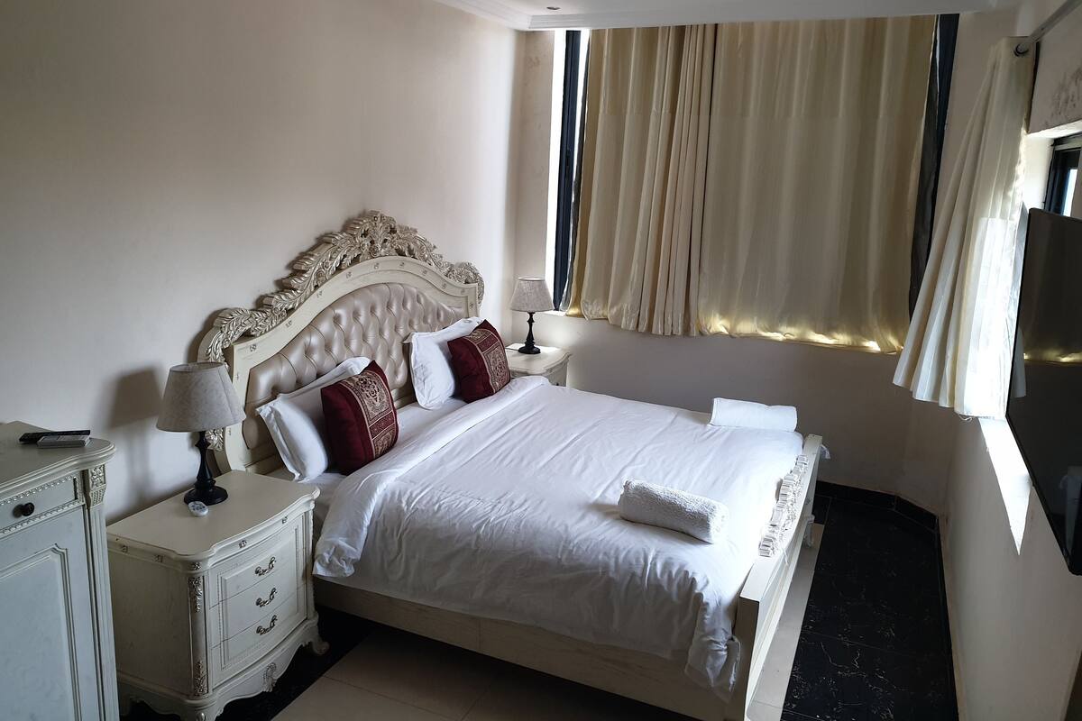 A neatly made bed with white linens and red pillows, a white nightstand with a lamp, and a dresser with drawers.