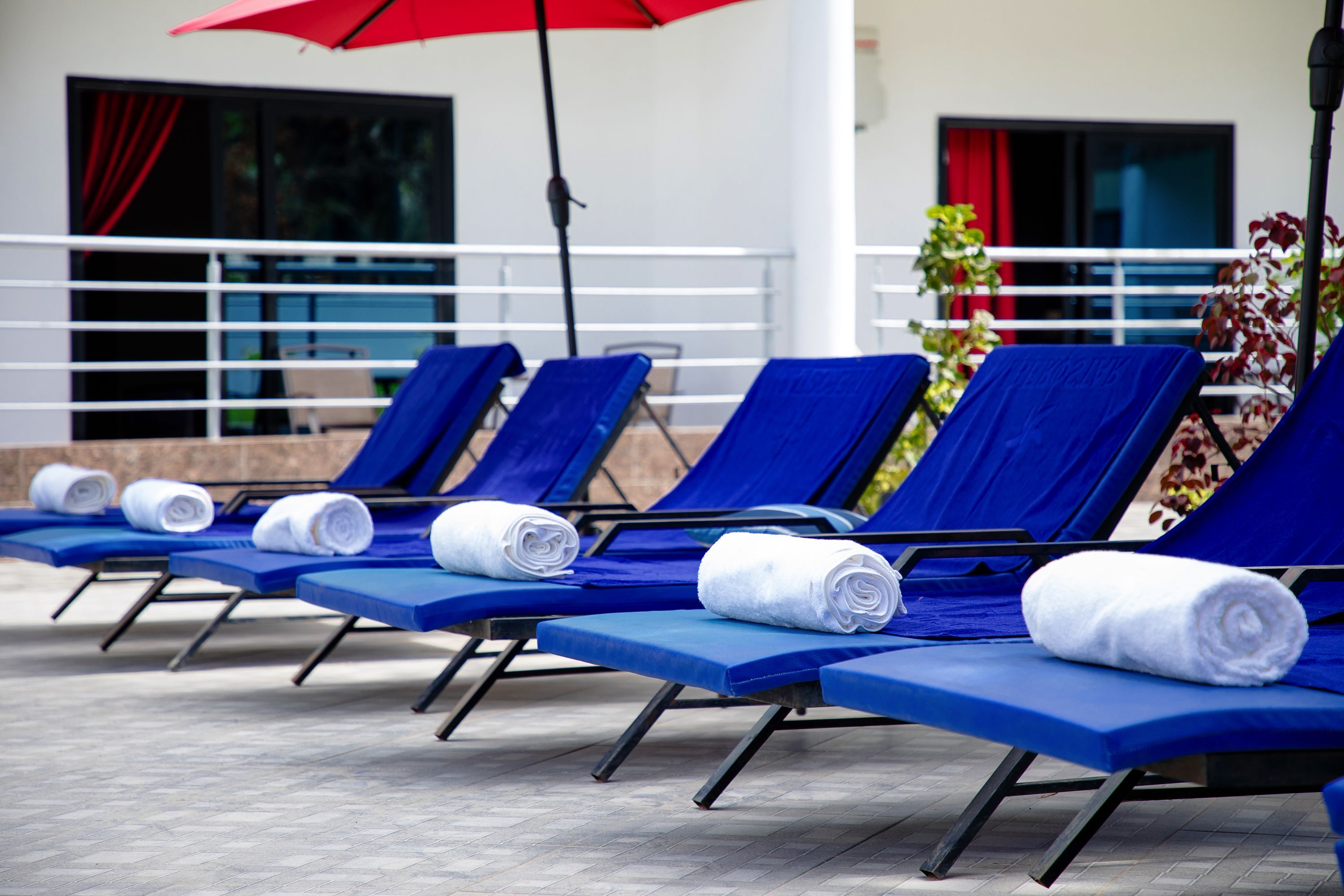 A row of blue lounge chairs with towels, a red umbrella, and a building with windows and a railing in the background.