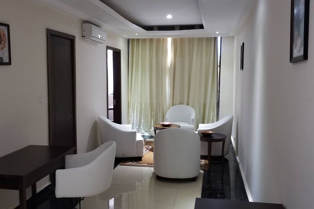 A modern hotel room with a dining area, a sofa, a small table, and a large window with curtains.
