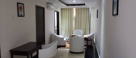 Una habitación de hotel moderna con zona de comedor, un sofá, una mesita y un ventanal grande con cortinas.