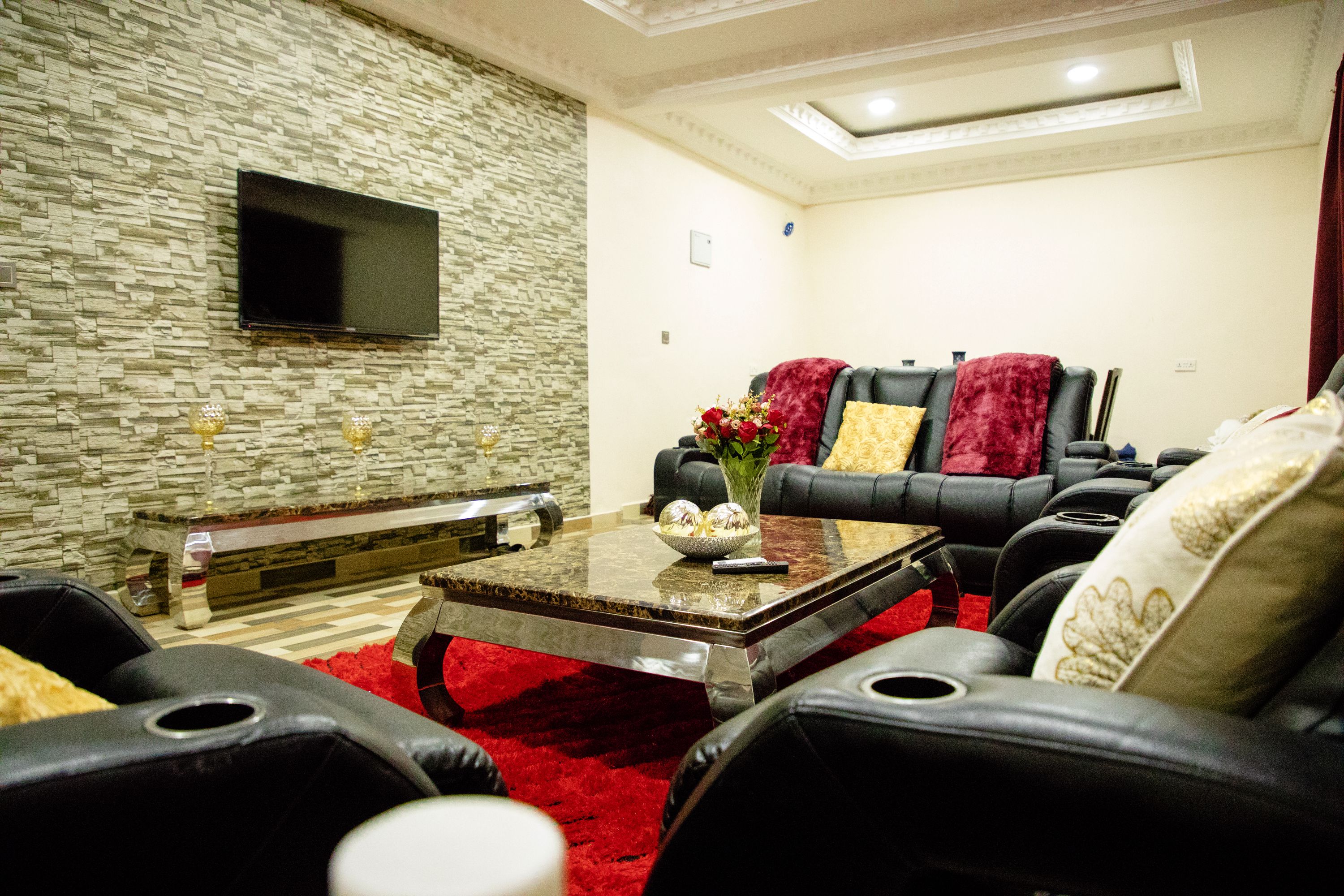 A modern living room with a stone wall, a flat-screen TV, a red carpet, and leather furniture.