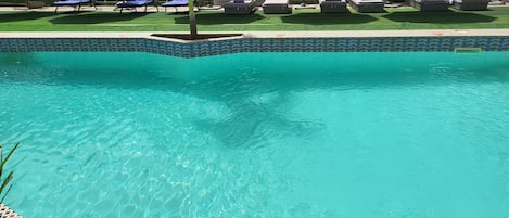 A swimming pool with clear blue water, surrounded by lounge chairs and a tiled deck.