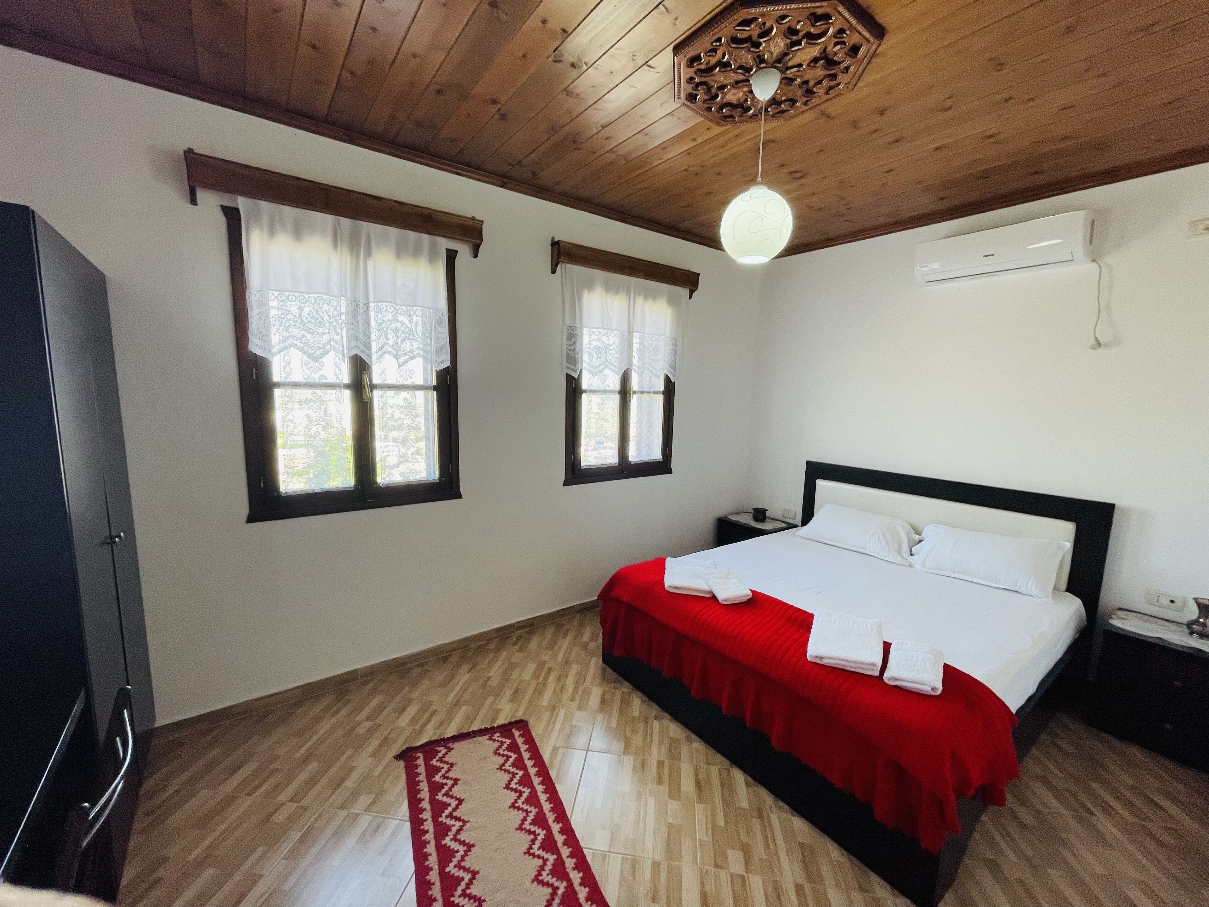 Ein Schlafzimmer mit Holzboden, zwei Fenstern mit Vorhängen, einem Bett mit roter Bettdecke, einem Nachttisch und einer Wandklimaanlage.