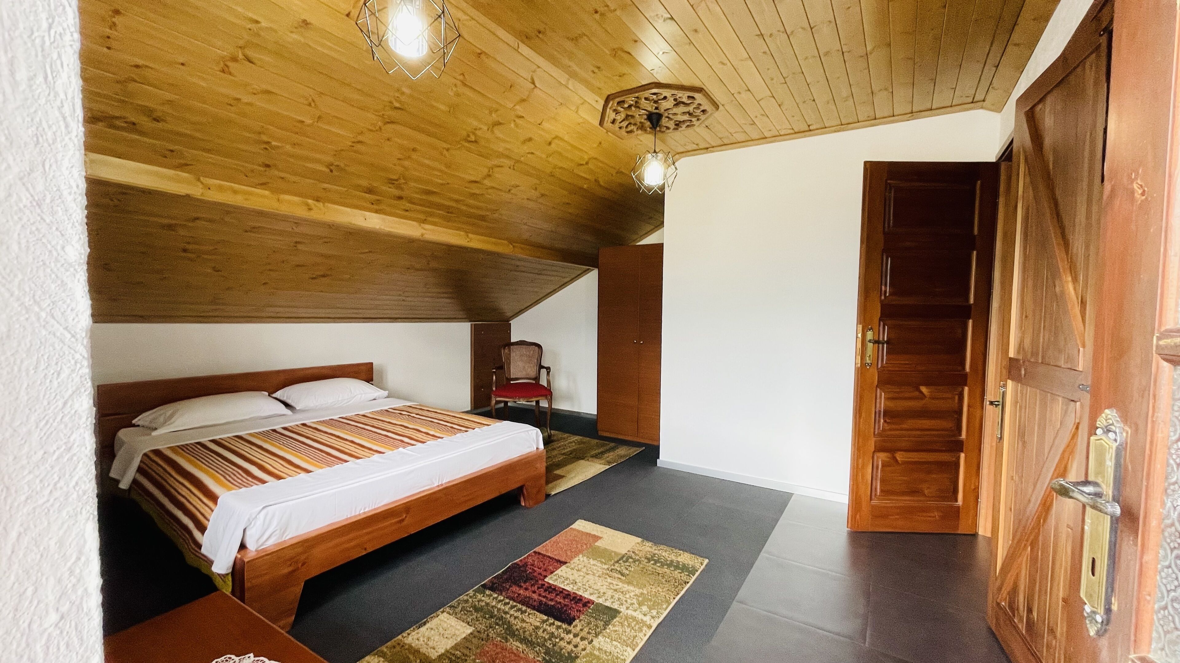 Un dormitorio con techo de madera, una cama con manta a rayas, una silla y un armario de madera.