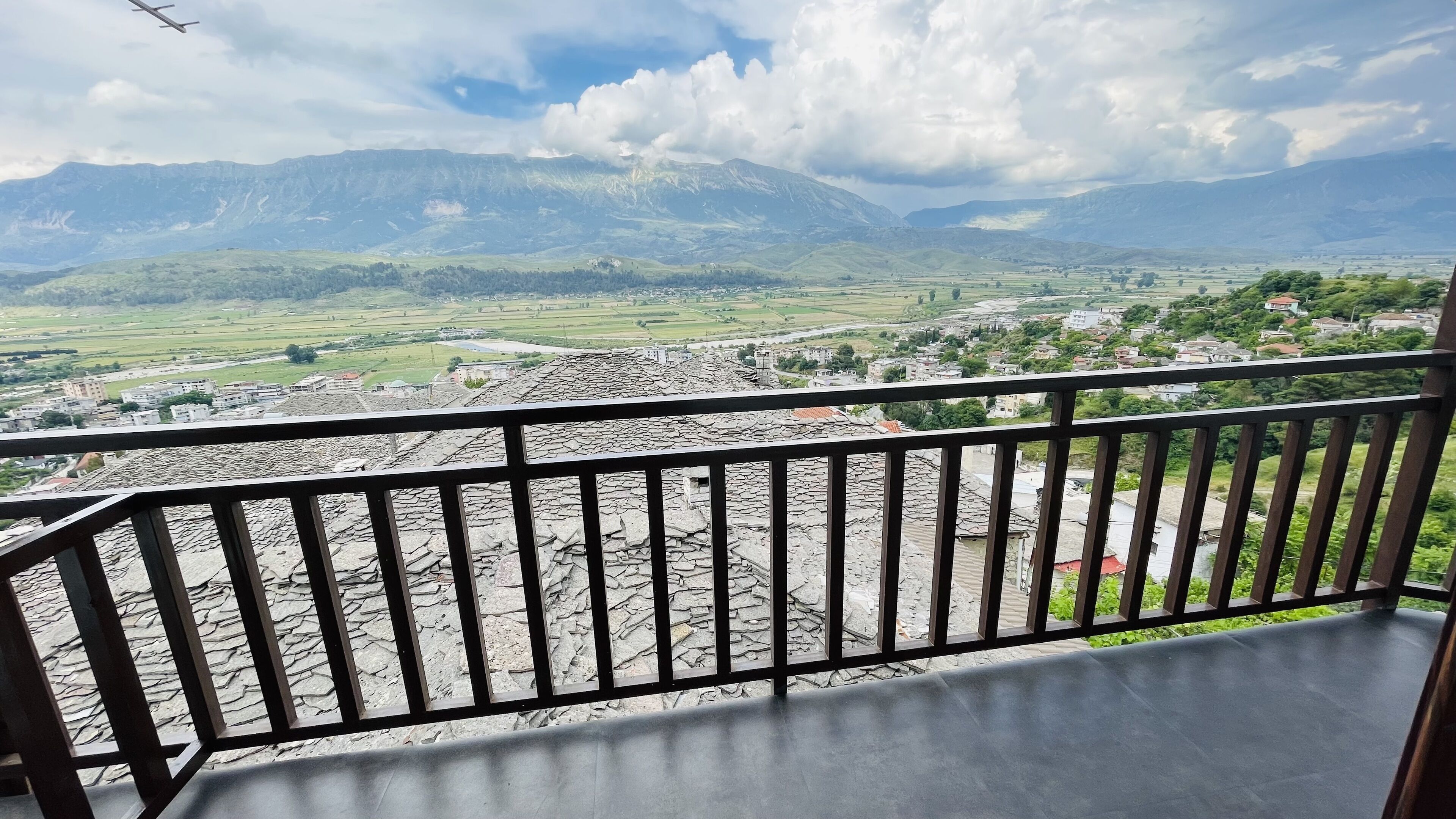 Un balcón con suelo de piedra y baranda de metal, con vistas a un valle con campos, un pueblo pequeño y montañas a la distancia.