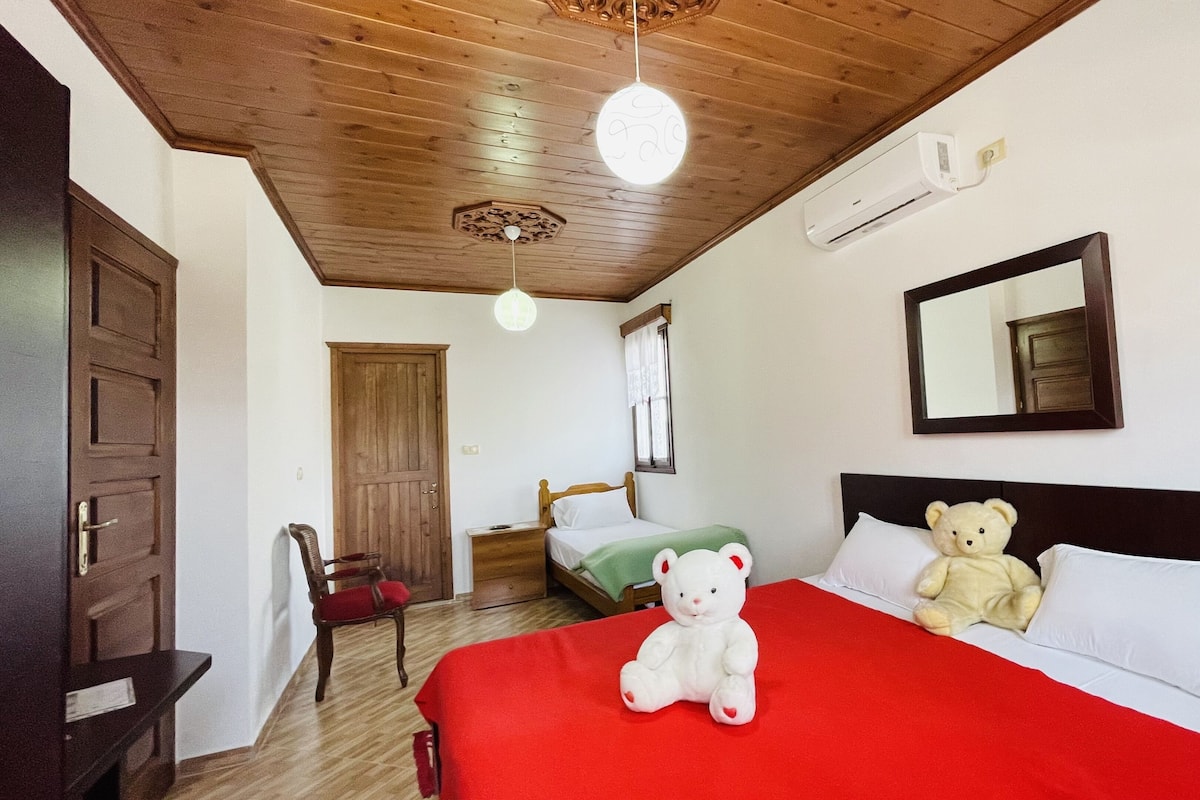Una habitación de hotel con techo de madera, una cama con manta roja, un oso de peluche blanco, una silla verde y un armario de madera.