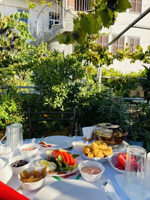 Mesa de comedor al aire libre con diversos platos, entre ellos frutas, verduras y condimentos, con un fondo de vegetación y un edificio.