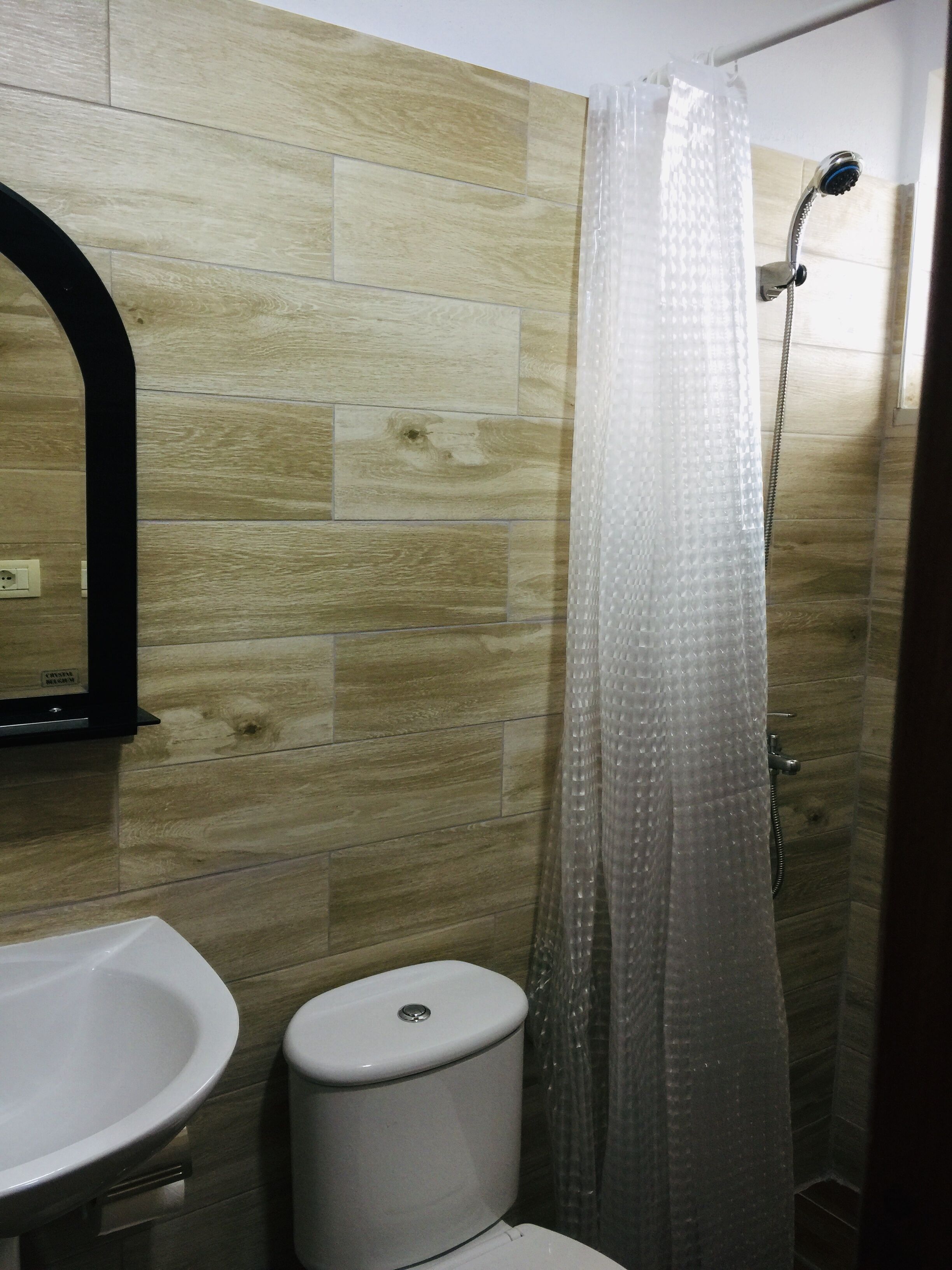 A bathroom with a wooden wall, a white sink, a toilet, and a shower with a transparent curtain.