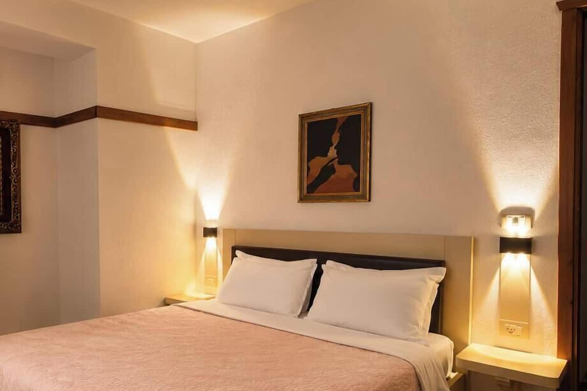 Una habitación de hotel con una cama grande, dos mesitas de noche, una lámpara de noche, un cuadro en la pared y luces fijadas a la pared.