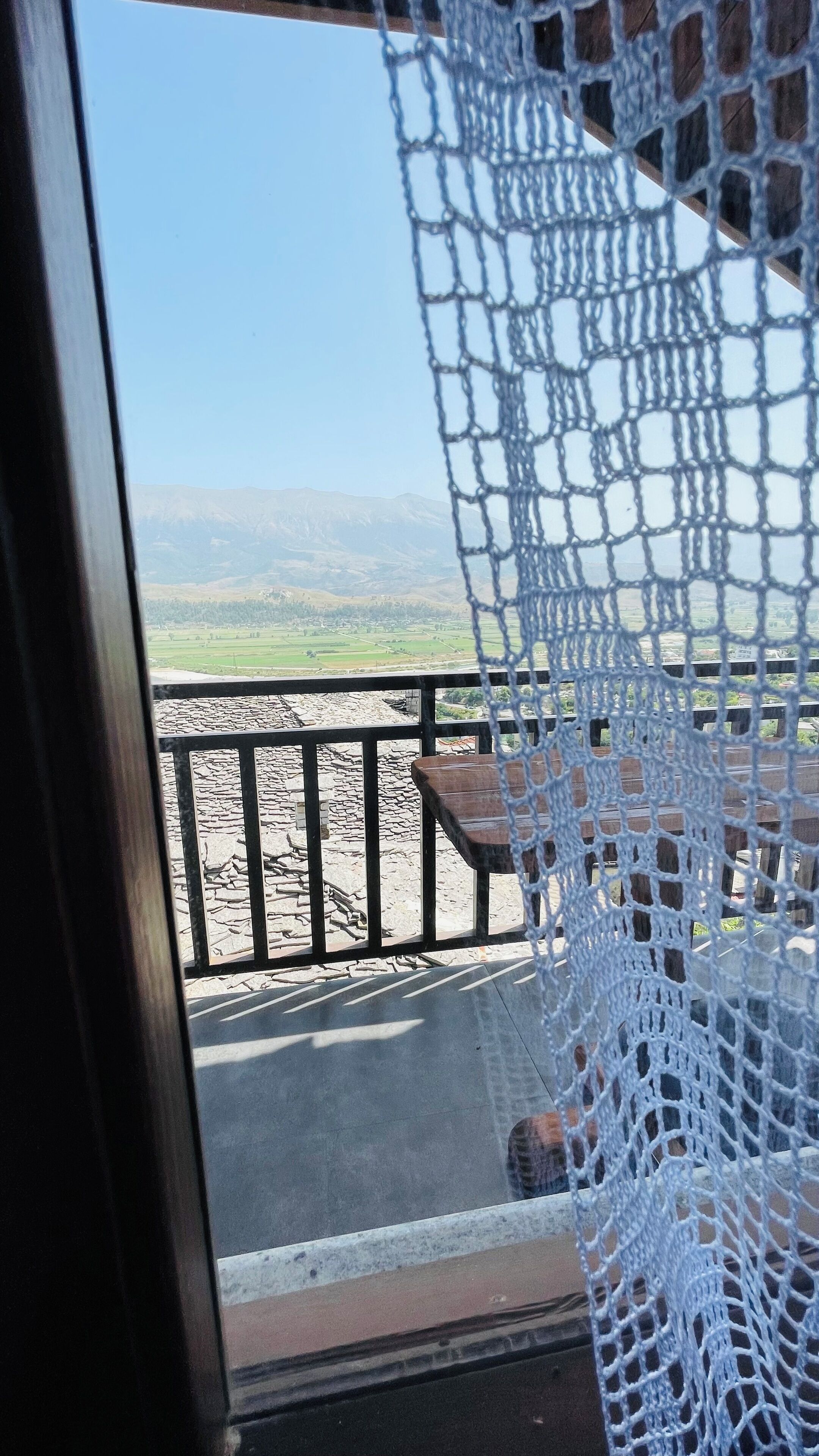 A view through a window with a white net, a stone floor, a wooden bench, and a stone railing.