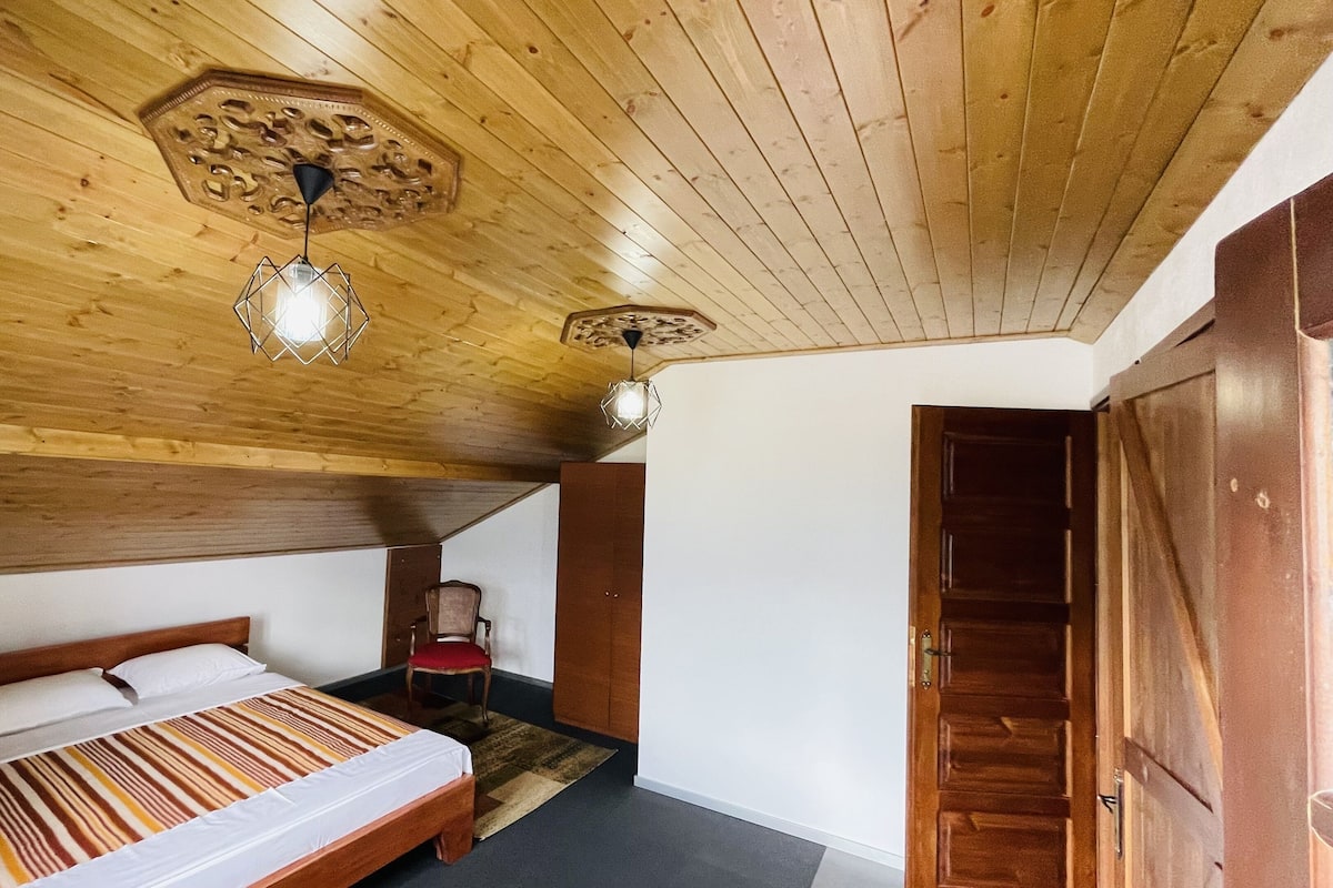 Techo de madera con dos lámparas colgantes, una cama con colcha a rayas, una silla y una puerta de madera.