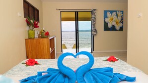 A hotel room with a bed, a desk with a vase of flowers, a painting of flowers, and a view of the sea through a sliding door.