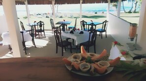 An outdoor dining area with tables and chairs, overlooking a beach. - Blue Bay Resort (Mele)