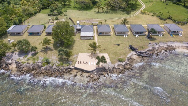 An aerial view of a resort with multiple bungalow-style buildings arranged in a row, a sandy beach area with footprints, and a clear view of the ocean.