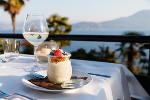 A dessert on a white table with a glass of white wine, set against a scenic view of the sea and mountains.